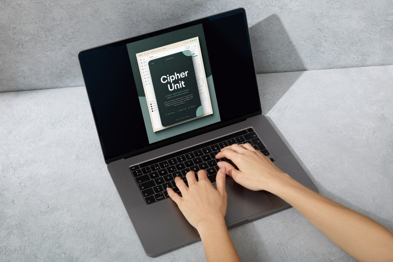 A top-down view of a person typing on a dark gray laptop with a screen displaying a book cover titled 'Cipher Unit', set against a textured gray concrete background.