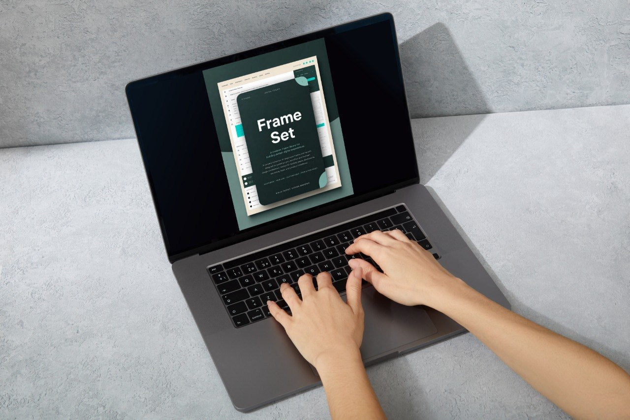 Top-down view of a person typing on a dark gray laptop computer, with the screen displaying a digital template or book cover titled 'Frame Set' against a textured gray background.
