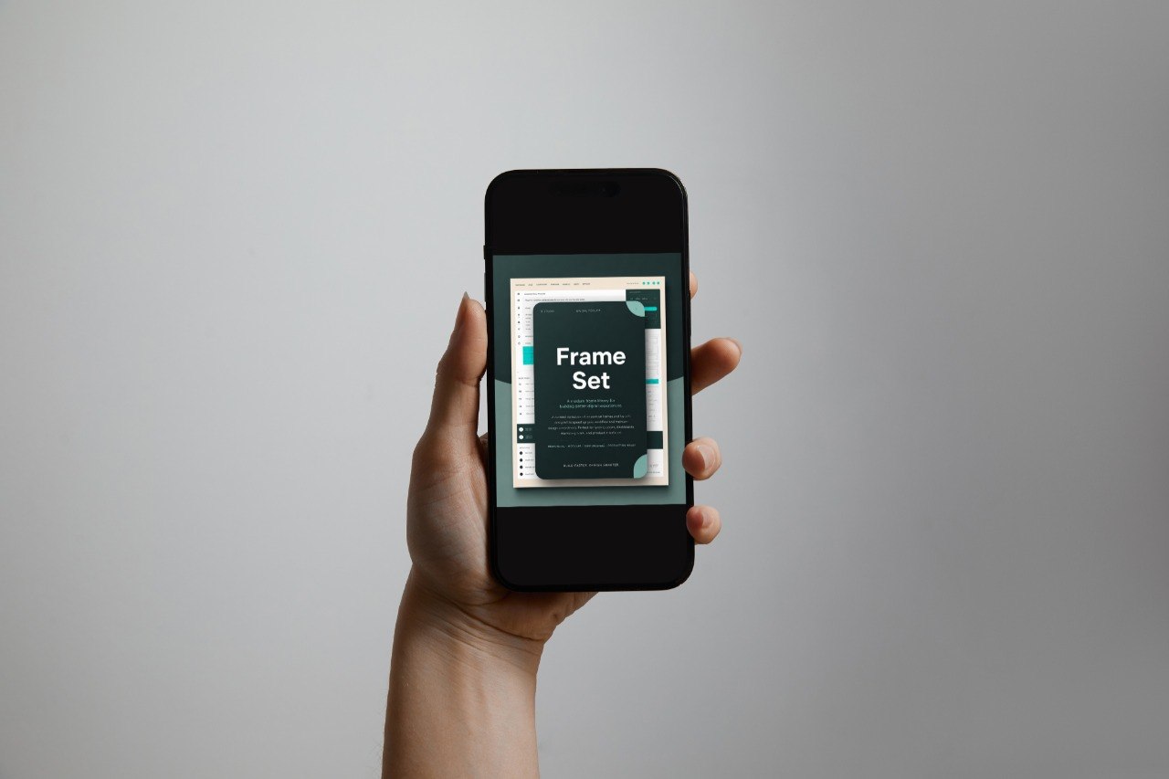 A hand holding a black smartphone against a plain light gray background. The phone screen displays a mockup or cover design with the text 'Frame Set'.